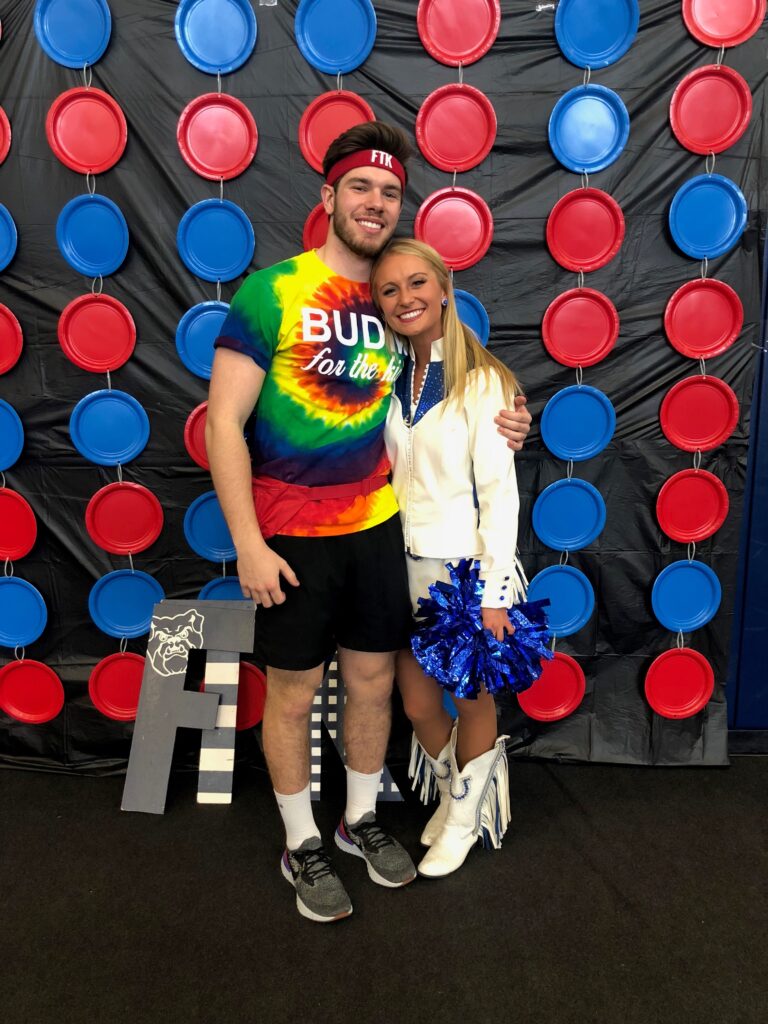 Nathan Jent wearing a colorful tie-dye BUDM t-shirt, standing next to Sarah Sharpe, wearing a white jacket and skirt, white cowboy boots, and holding a blue cheerleading pom pom.